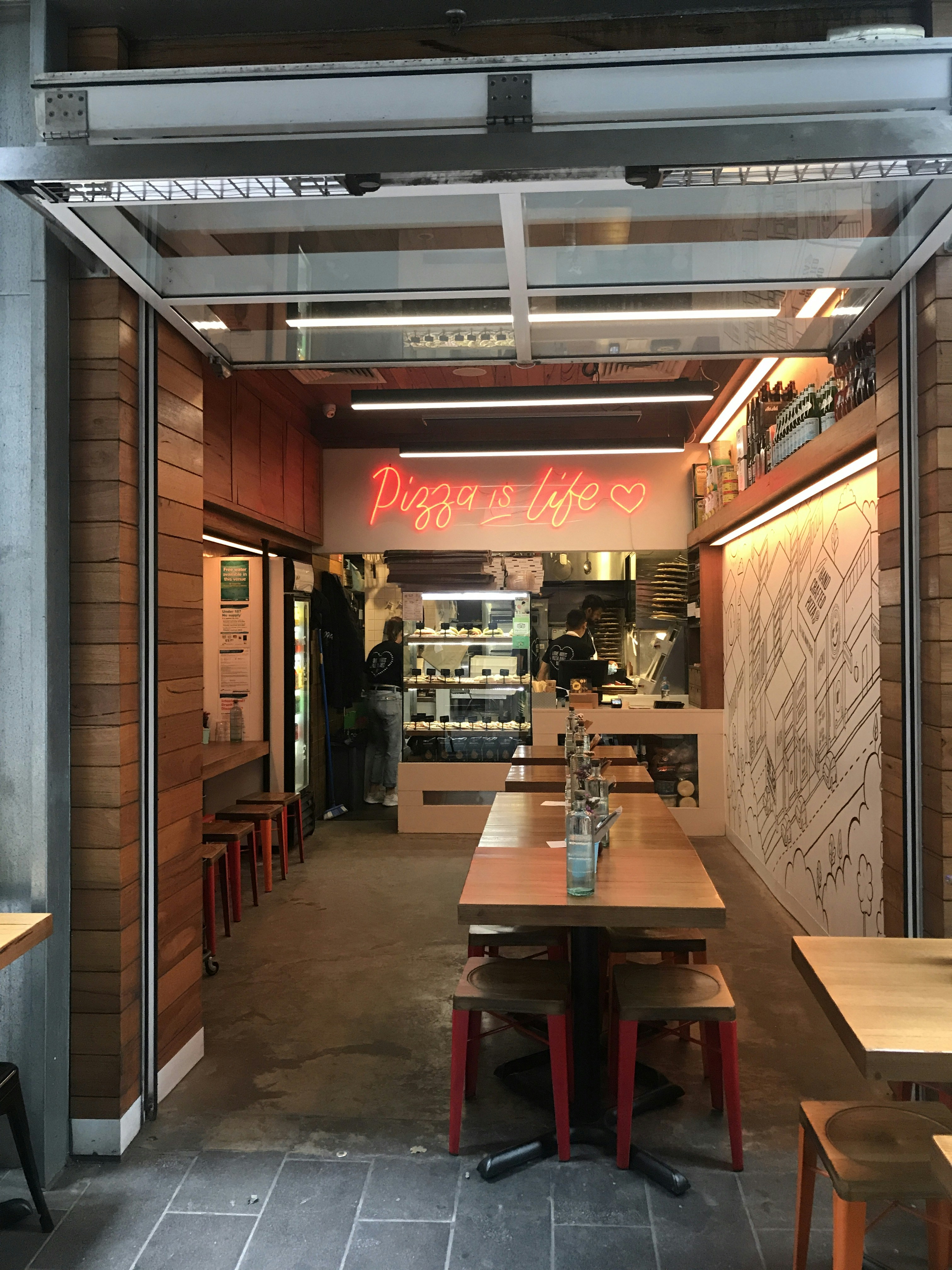A cozy pizza restaurant with an open entrance, featuring wooden tables and red stools. Inside, there is a neon sign with the phrase 'Pizza is life' and a heart symbol. The interior displays a counter with pizza ovens and a wall with artistic line drawings. Shelves with bottled beverages line one side of the establishment.