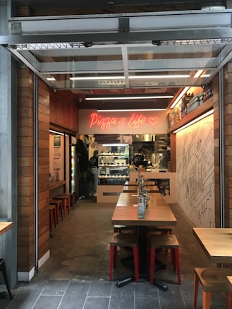 A cozy pizza restaurant with an open entrance, featuring wooden tables and red stools. Inside, there is a neon sign with the phrase 'Pizza is life' and a heart symbol. The interior displays a counter with pizza ovens and a wall with artistic line drawings. Shelves with bottled beverages line one side of the establishment.