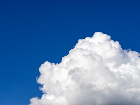 A large, fluffy white cloud against a bright blue sky, creating a striking contrast. The cloud has a billowy appearance with soft edges and varying shades of white and gray, indicating depth and texture.