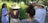 A group of people gathers outdoors in a lush garden, listening intently to a woman with long hair, who is speaking and gesturing. Sunlight filters through the trees, casting dappled shadows on the group. Everyone wears casual summer clothing, with several wide-brimmed hats providing shade from the sun.