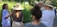 A group of people gathers outdoors in a lush garden, listening intently to a woman with long hair, who is speaking and gesturing. Sunlight filters through the trees, casting dappled shadows on the group. Everyone wears casual summer clothing, with several wide-brimmed hats providing shade from the sun.