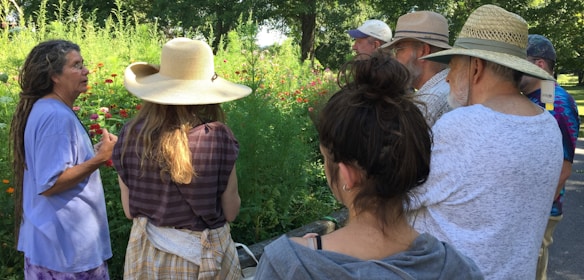A group of people gathers outdoors in a lush garden, listening intently to a woman with long hair, who is speaking and gesturing. Sunlight filters through the trees, casting dappled shadows on the group. Everyone wears casual summer clothing, with several wide-brimmed hats providing shade from the sun.