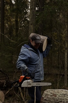 A person in a forest setting carries a log on their shoulder while holding a chainsaw. The individual is wearing a blue jacket and a cap, surrounded by tall trees and natural foliage.
