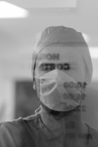 A medical professional is seen wearing a surgical mask and cap, with a reflective surface showing blurred text in the foreground. The image is captured in black and white, giving it a classic and serious tone.