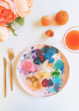 A vibrant tabletop composition featuring a painter's palette with an array of colorful paints. Next to the palette are a few pieces of fruit, including an orange, a peach, and a strawberry. A glass with an orange-hued liquid is on the right, while a gold fork and knife are placed on the left. Peonies in soft pink shades add a floral touch to the scene.