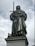 A metal statue of a regal figure stands atop a stone pedestal, holding a sword in an upright position. The figure is dressed in flowing robes and a crown, exuding an aura of authority and strength.