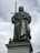A metal statue of a regal figure stands atop a stone pedestal, holding a sword in an upright position. The figure is dressed in flowing robes and a crown, exuding an aura of authority and strength.