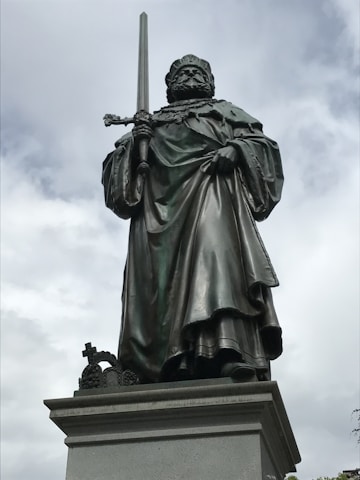 A metal statue of a regal figure stands atop a stone pedestal, holding a sword in an upright position. The figure is dressed in flowing robes and a crown, exuding an aura of authority and strength.