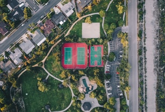 An aerial view of a residential area with neatly arranged houses along a road. Adjacent to the houses, there is a park featuring tennis and basketball courts with bright markings. The park area is surrounded by pathways and scattered trees, providing some greenery amongst the urban setting. To the right, there is a parking lot with multiple cars parked, and a road running parallel to it.