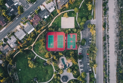 An aerial view of a residential area with neatly arranged houses along a road. Adjacent to the houses, there is a park featuring tennis and basketball courts with bright markings. The park area is surrounded by pathways and scattered trees, providing some greenery amongst the urban setting. To the right, there is a parking lot with multiple cars parked, and a road running parallel to it.