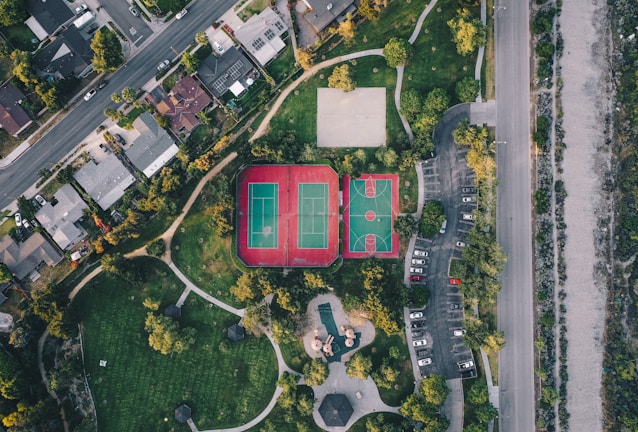 An aerial view of a residential area with neatly arranged houses along a road. Adjacent to the houses, there is a park featuring tennis and basketball courts with bright markings. The park area is surrounded by pathways and scattered trees, providing some greenery amongst the urban setting. To the right, there is a parking lot with multiple cars parked, and a road running parallel to it.