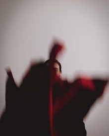 A partially obscured person is standing against a plain, light-colored background. A blurred fabric with hints of red is in the foreground, adding an artistic and abstract element to the composition.