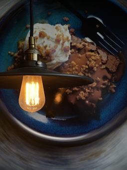 A dessert plate features a slice of chocolate cake topped with caramel sauce and crumbled nuts, accompanied by a scoop of vanilla ice cream. An Edison bulb hanging light casts a warm glow over the dessert, creating an inviting and cozy atmosphere.