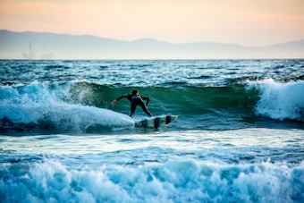 A surfer rides a wave in an ocean setting, with the sun setting behind distant mountains. The water is animated and dynamic, while the surfer skillfully navigates through the foamy waves.