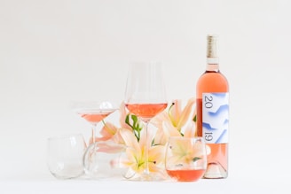 A still life arrangement featuring a bottle of pink rosé wine with a label displaying the year 2019. Several glasses, some filled with the wine, are positioned artistically around the bottle, alongside soft pink lilies. The composition rests on a neutral background, emphasizing the elegance of the flowers and glassware.