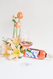 A bouquet of orange and peach flowers is arranged gracefully next to a bottle of rosé wine, which lays on a flat surface. A glass of the pink wine is partially filled, standing elegantly in the foreground. The background is a soft, neutral color.