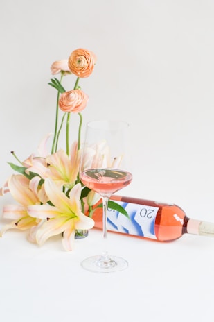 A bouquet of orange and peach flowers is arranged gracefully next to a bottle of rosé wine, which lays on a flat surface. A glass of the pink wine is partially filled, standing elegantly in the foreground. The background is a soft, neutral color.