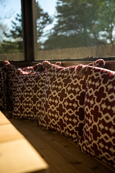 A row of decorative cushions with a geometric pattern in dark red and white are placed along a wooden bench by a large window. The sunlight streams through the window, creating a warm ambiance, and the view outside shows blurred trees and a clear sky.