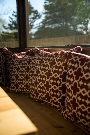 A row of decorative cushions with a geometric pattern in dark red and white are placed along a wooden bench by a large window. The sunlight streams through the window, creating a warm ambiance, and the view outside shows blurred trees and a clear sky.