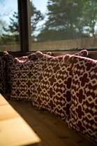 A row of decorative cushions with a geometric pattern in dark red and white are placed along a wooden bench by a large window. The sunlight streams through the window, creating a warm ambiance, and the view outside shows blurred trees and a clear sky.