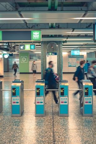 A subway or train station entrance area with several people using turnstiles. Signage with directions and exits is visible above, including a prominent green 'EXIT' sign. The floor and walls are tiled, and the environment appears clean and organized.