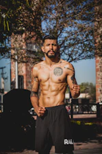 A shirtless man with tattoos on his chest and arms stands outdoors, with a confident and focused expression, set against a background of trees and brick structures. He has a well-defined muscular build and is wearing black athletic shorts.