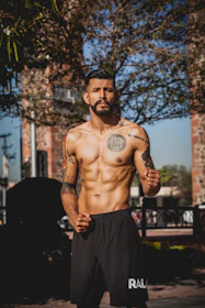 A shirtless man with tattoos on his chest and arms stands outdoors, with a confident and focused expression, set against a background of trees and brick structures. He has a well-defined muscular build and is wearing black athletic shorts.