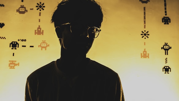 A silhouette of a person is set against a background filled with pixelated, retro-style video game graphics. The lighting creates a dramatic contrast, emphasizing the outline of the person, who is wearing glasses. The graphics include various pixelated characters and symbols that evoke a classic arcade game theme.