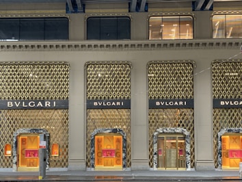 A luxury storefront with a lavish gold-accented facade, featuring prominent branding of the name 'BVLGARI'. The store displays have illuminated windows showcasing products, with elegant design and decor elements. The overall aesthetic conveys sophistication and opulence.