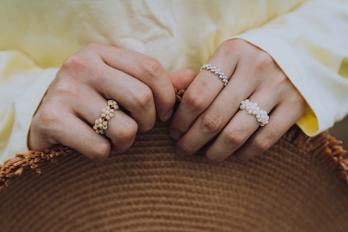 Hands with several beaded rings are gently holding a textured straw hat. The rings feature small, delicate beads in white and neutral tones. The person's shirt sleeves are light yellow, softly blending in with the overall earthy color palette.