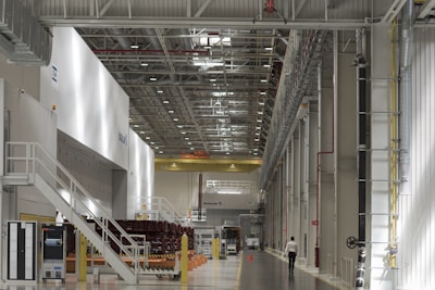 A spacious industrial facility with high ceilings and extensive use of metal and industrial materials. The interior is well-lit, featuring clean, organized rows of machinery and equipment. A single person is walking in the distance, emphasizing the scale of the space. The environment appears to be a manufacturing or industrial warehouse with marked walkways and large doors.