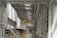 A spacious industrial facility with high ceilings and extensive use of metal and industrial materials. The interior is well-lit, featuring clean, organized rows of machinery and equipment. A single person is walking in the distance, emphasizing the scale of the space. The environment appears to be a manufacturing or industrial warehouse with marked walkways and large doors.