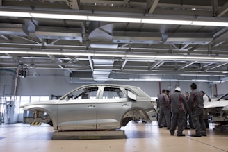 A car body is positioned on a platform in a spacious, well-lit factory or workshop. The metallic framework of the vehicle is accompanied by a group of workers clad in matching uniforms, likely discussing or inspecting the vehicle. Overhead, there are large metal beams and lighting fixtures, indicating an industrial setting.