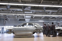 A car body is positioned on a platform in a spacious, well-lit factory or workshop. The metallic framework of the vehicle is accompanied by a group of workers clad in matching uniforms, likely discussing or inspecting the vehicle. Overhead, there are large metal beams and lighting fixtures, indicating an industrial setting.