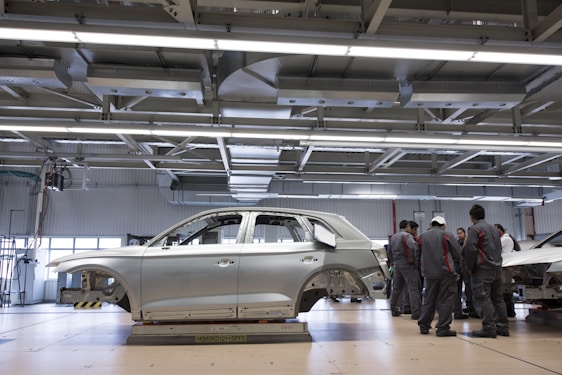 A car body is positioned on a platform in a spacious, well-lit factory or workshop. The metallic framework of the vehicle is accompanied by a group of workers clad in matching uniforms, likely discussing or inspecting the vehicle. Overhead, there are large metal beams and lighting fixtures, indicating an industrial setting.