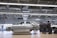 A car body is positioned on a platform in a spacious, well-lit factory or workshop. The metallic framework of the vehicle is accompanied by a group of workers clad in matching uniforms, likely discussing or inspecting the vehicle. Overhead, there are large metal beams and lighting fixtures, indicating an industrial setting.