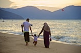 A family of three is walking along a sandy beach with gentle waves rolling in. The parents are holding hands with their child as they stroll. In the background, hills are silhouetted against a sunset sky. The atmosphere is peaceful and serene.