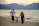 A family of three is walking along a sandy beach with gentle waves rolling in. The parents are holding hands with their child as they stroll. In the background, hills are silhouetted against a sunset sky. The atmosphere is peaceful and serene.