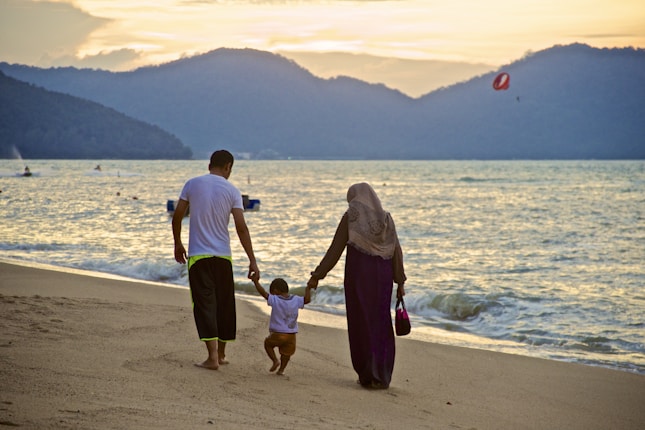 A family of three is walking along a sandy beach with gentle waves rolling in. The parents are holding hands with their child as they stroll. In the background, hills are silhouetted against a sunset sky. The atmosphere is peaceful and serene.