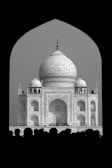 The image features a monochrome view of the Taj Mahal framed within a dark archway. The grand architecture, with its large dome and intricate details, stands out against the lighter background. Silhouettes of people are visible at the bottom, adding a sense of scale and human presence.