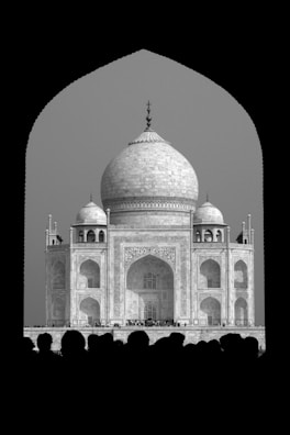 The image features a monochrome view of the Taj Mahal framed within a dark archway. The grand architecture, with its large dome and intricate details, stands out against the lighter background. Silhouettes of people are visible at the bottom, adding a sense of scale and human presence.