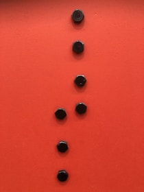Seven black hexagonal bolts are arranged vertically on a bright orange surface. The bolts are evenly spaced and create a linear pattern against the vibrant background.