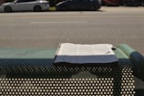 A newspaper is placed on a perforated metal bench by the side of a road. In the background, there are several parked cars along a wide street. Shadows from the bench and the newspaper are visible on the ground.