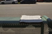 A newspaper is placed on a perforated metal bench by the side of a road. In the background, there are several parked cars along a wide street. Shadows from the bench and the newspaper are visible on the ground.
