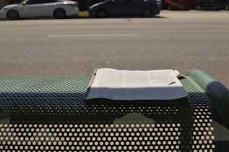 A newspaper is placed on a perforated metal bench by the side of a road. In the background, there are several parked cars along a wide street. Shadows from the bench and the newspaper are visible on the ground.
