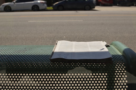 A newspaper is placed on a perforated metal bench by the side of a road. In the background, there are several parked cars along a wide street. Shadows from the bench and the newspaper are visible on the ground.