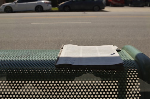 A newspaper is placed on a perforated metal bench by the side of a road. In the background, there are several parked cars along a wide street. Shadows from the bench and the newspaper are visible on the ground.