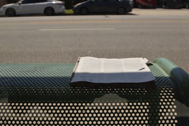 A newspaper is placed on a perforated metal bench by the side of a road. In the background, there are several parked cars along a wide street. Shadows from the bench and the newspaper are visible on the ground.