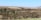 Rolling hills covered with dry grass and scattered trees create a tranquil landscape. The mix of brown and green earth tones blend under a clear blue sky.