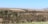 Rolling hills covered with dry grass and scattered trees create a tranquil landscape. The mix of brown and green earth tones blend under a clear blue sky.